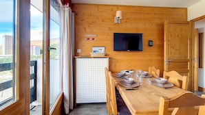 Ein Zimmer mit Holzwänden, einem Esstisch für vier Personen, einem an der Wand montierten Fernseher und Blick auf die Stadt durch große Fenster.