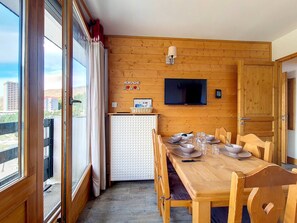 Ein Zimmer mit Holzwänden, einem Esstisch für vier Personen, einem an der Wand montierten Fernseher und Blick auf die Stadt durch große Fenster.