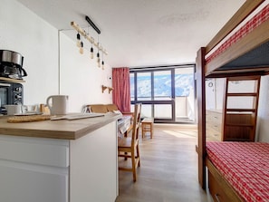 Cozinha compacta com cafeteira, área de refeições com mesa e cadeiras, beliche com edredom xadrez vermelho e uma grande janela com vista para as montanhas.