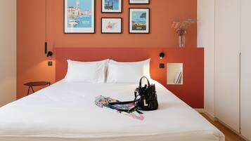 A neatly made bed with white linens, a black handbag, and a colorful scarf on the bed. There are framed pictures on the wall above the bed.