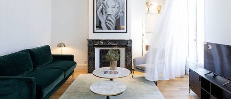 A modern living room with a dark green sofa, a round marble coffee table, a fireplace, and a large window with sheer curtains.