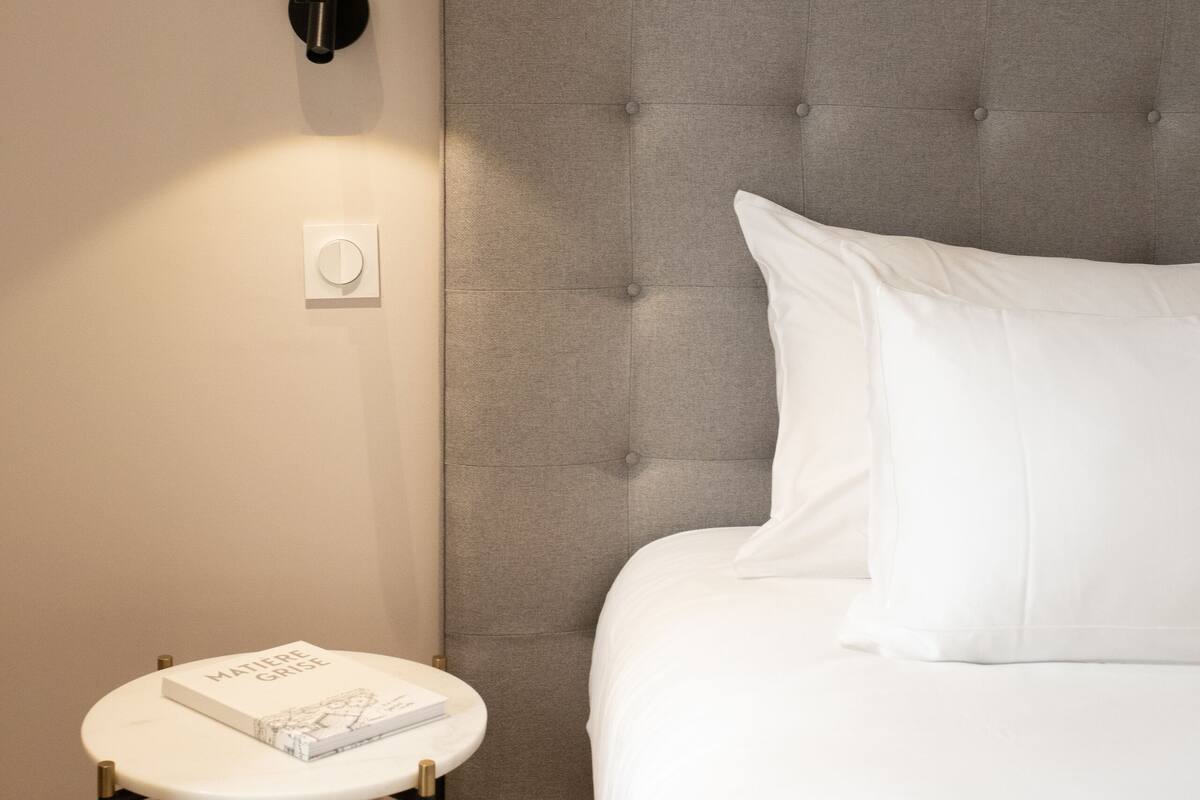 Una cama bien hecha con un cabecero gris con capitoné, una mesita de noche blanca con un libro y una lámpara fijada a la pared.
