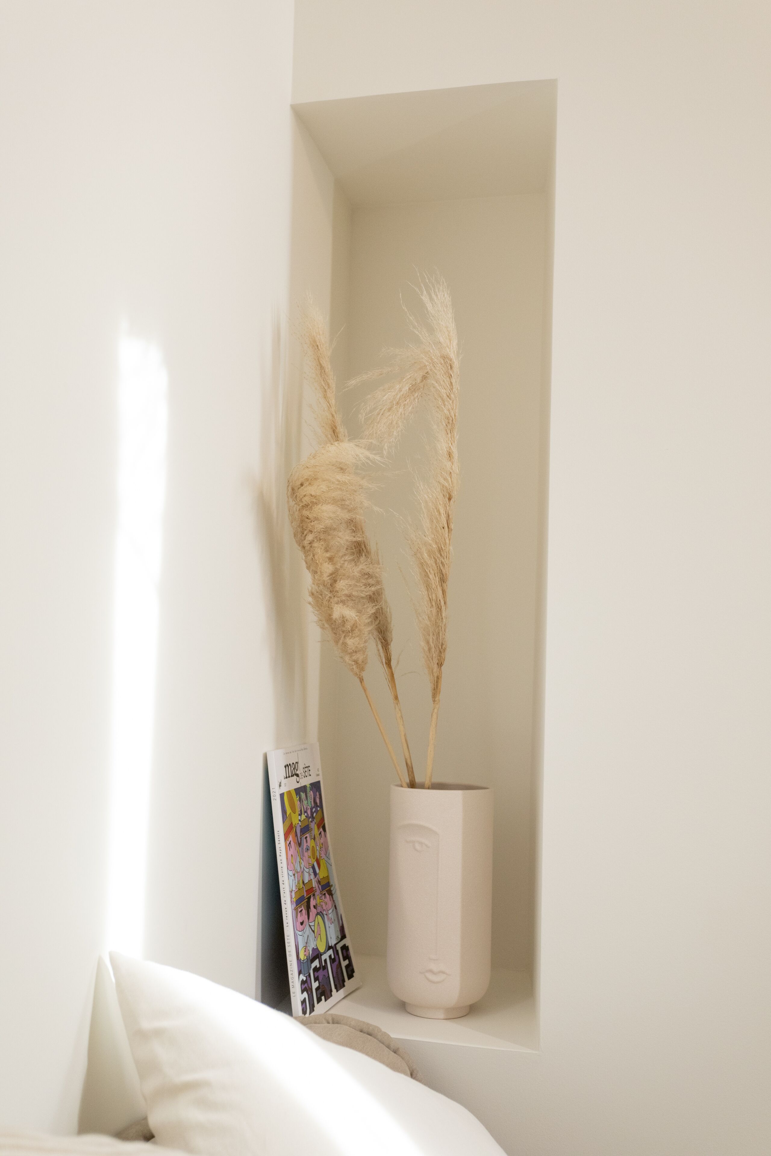 Eine weiße Wand mit einem kleinen Regal, auf dem sich eine weiße Vase mit Pampasgras und eine Zeitschrift befinden.