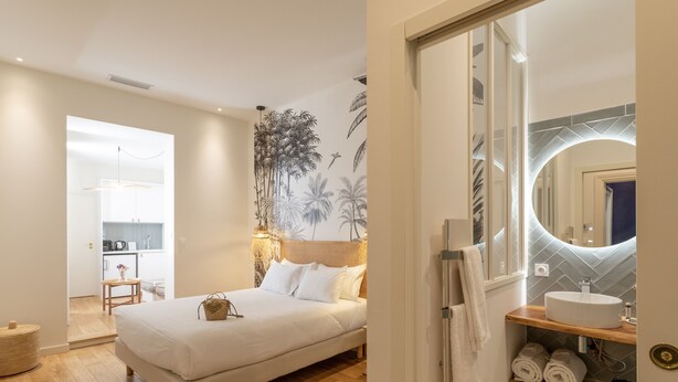 A bedroom with a bed, a bedside table, a wall-mounted mirror, and a bathroom with a sink and towel rack. - Les Clés Secrètes (Sète)