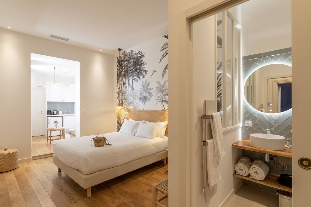 Un dormitorio con cama, una mesita de noche, un espejo montado en la pared y un baño con lavamanos y toallero.