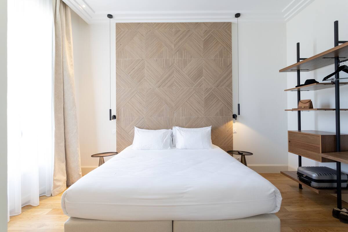 Habitación de hotel ordenada con una cama grande, cabecera de madera, armario con estantes y una mesita.