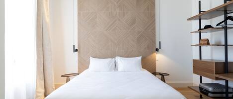 A neatly arranged hotel room with a large bed, a wooden headboard, a wardrobe with shelves, and a small table.