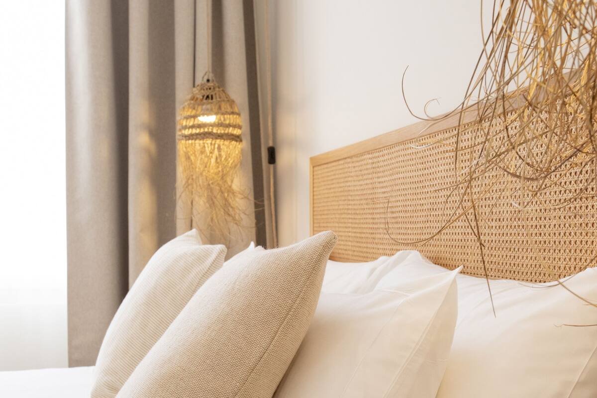 Una cama bien tendida con sábanas y almohadas blancas, un cabecero tejido y una lámpara decorativa.
