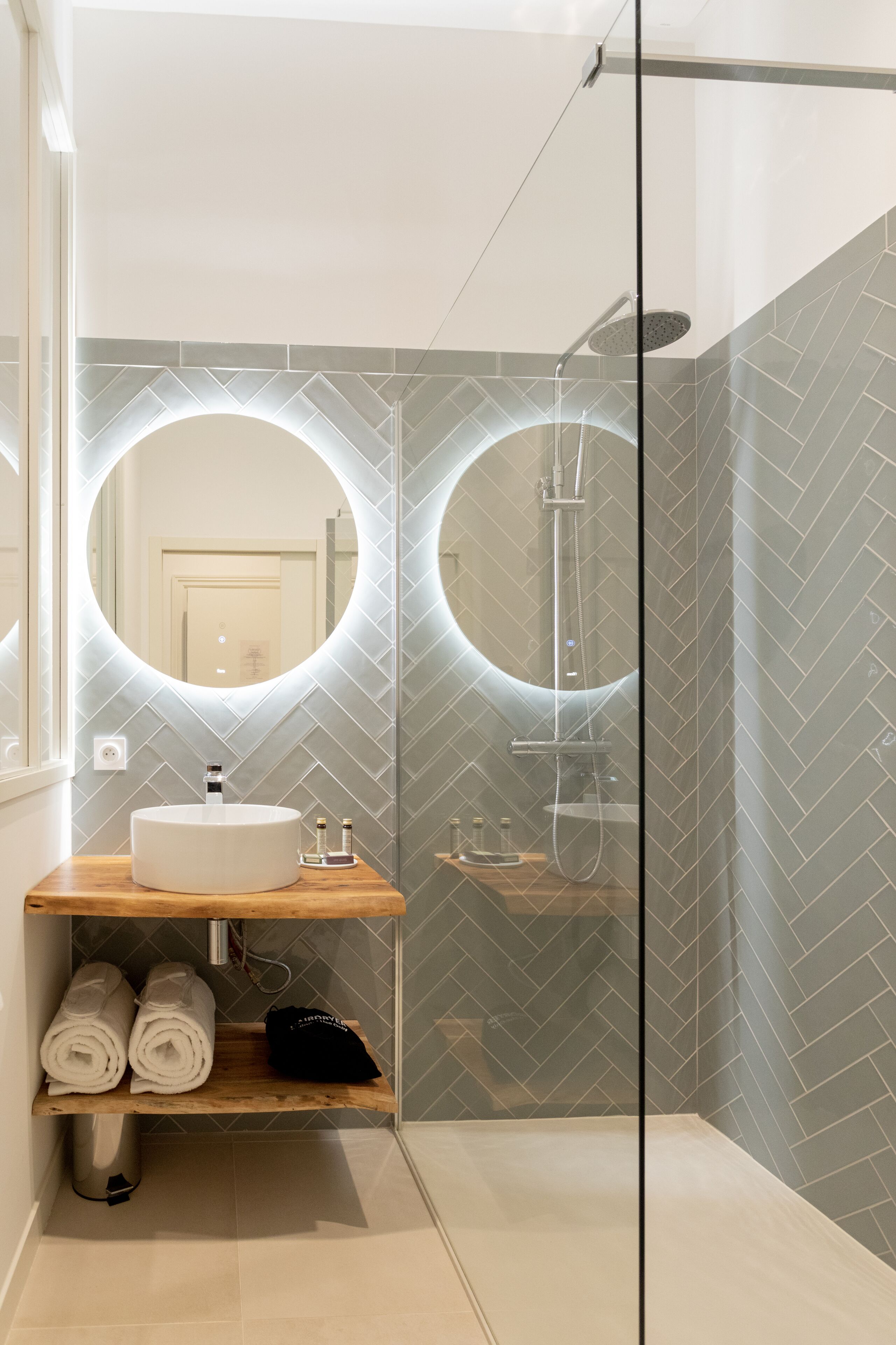 A modern bathroom with a glass-enclosed shower, a round mirror, and a wooden vanity with towels.