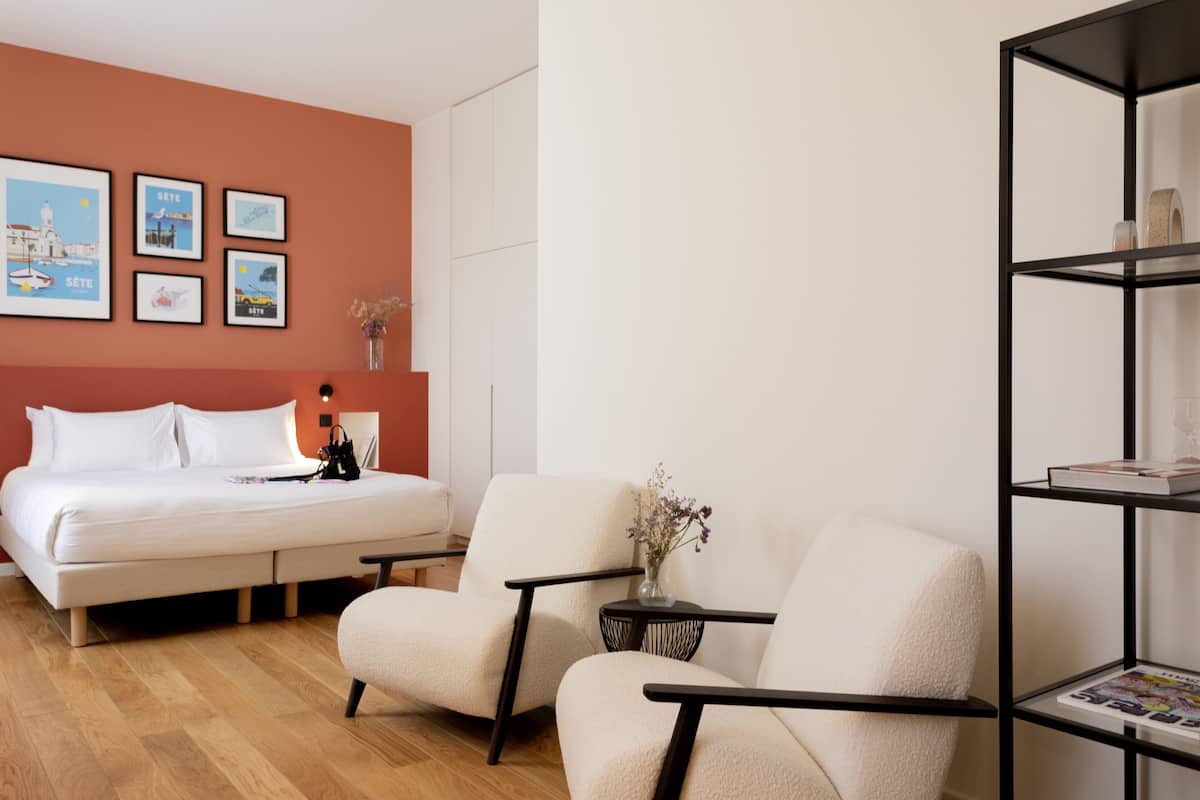 Una habitación de hotel moderna con una cama, dos sillones, una mesita con un florero y una estantería.