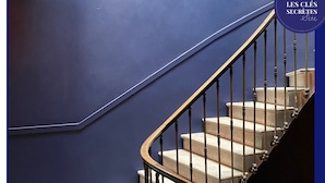A staircase with a dark blue wall and a wooden handrail. - Les Clés Secrètes (Sète)