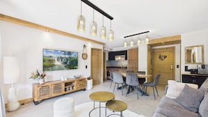 A modern living room with a sofa, coffee table, and dining area with chairs. There is a kitchen in the background with wooden cabinets and a dining table.