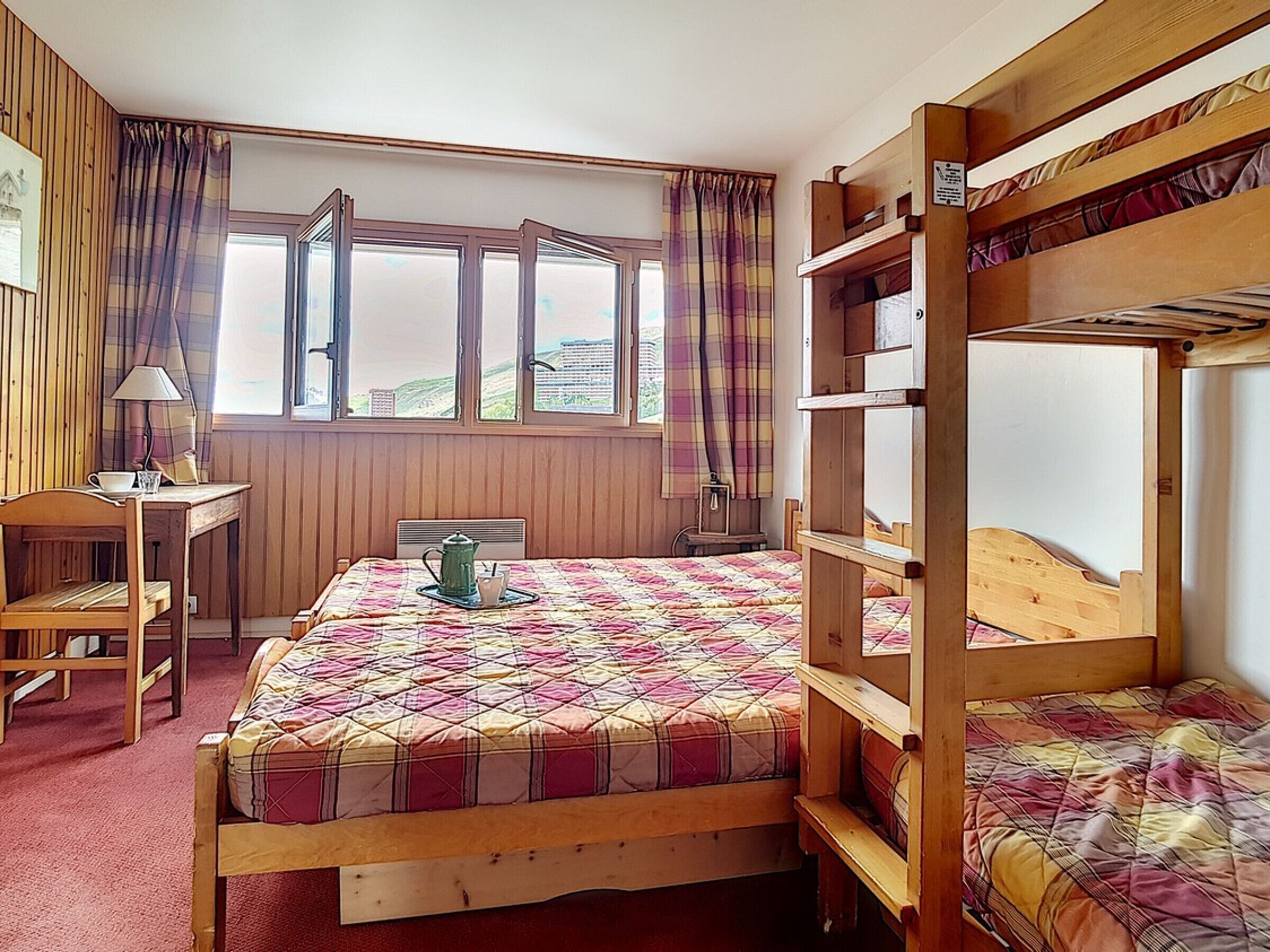 Une chambre avec un lit superposé, un bureau avec une lampe, une fenêtre avec des rideaux et un lit avec une couette.