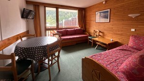 Una habitación acogedora con pared de madera, una cama con colcha estampada, una mesa de comedor con sillas, un sofá con cojines, un televisor montado en la pared y una ventana con cortinas.