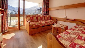 Una habitació acollidora amb un llit de fusta, un sofà amb una manta estampada, una taula petita amb un aparell de ràdio i una vista de muntanyes a través d'una finestra gran.