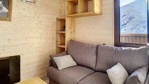 Interior acolhedor de uma cabana de madeira, com um sofá cinza, uma pequena estante de madeira e uma janela com vista para uma montanha coberta de neve.