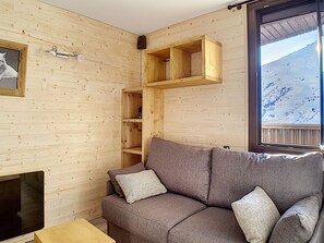 A cozy wooden cabin interior with a gray sofa, a small wooden shelf unit, and a window with a snowy mountain view.