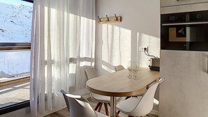 Uma área de refeições moderna com mesa de madeira e cadeiras brancas, uma cozinha com forno embutido, e vista para o exterior através de cortinas leves.