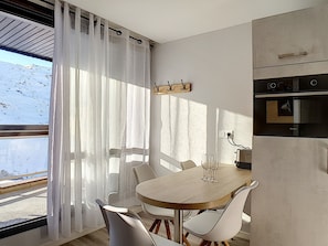 A modern dining area with a wooden table and white chairs, a kitchen with a built-in oven, and a view of the outdoors through sheer curtains.