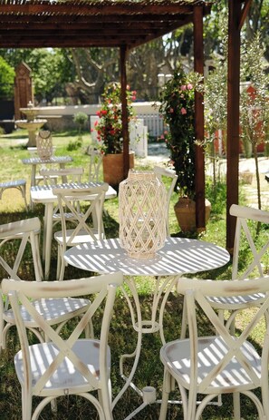 Outdoor seating with white metal chairs and tables, surrounded by greenery and a wooden pergola.