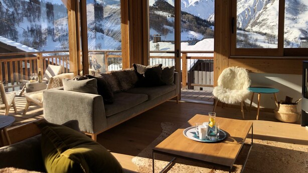 Una cabaña acogedora con interior de madera, grandes ventanales con vistas a las montañas, un sofá, sillas y una mesa con una bebida.