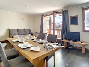 Ein modernes Wohnzimmer mit einem Holzespeisetisch, gedeckt für vier Personen, einer Couch, einem Fernseher und einem Balkon mit Blick auf eine verschneite Landschaft.