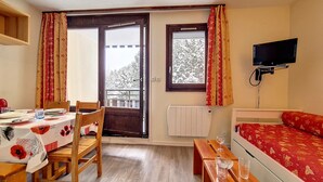 A room with a dining table set for two, a wooden bench, a bed with a red patterned bedspread, a TV mounted on the wall, and a window with curtains. - Cozy studio at the foot of the slopes, balcony, pets allowed (Les Menuires)