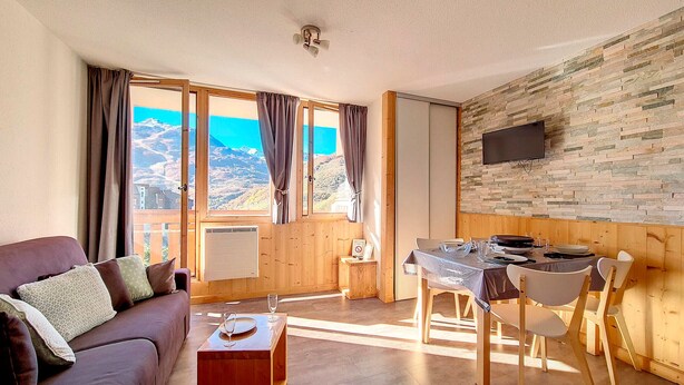 Uma sala de estar moderna com piso de madeira, um sofá marrom, uma mesa de jantar com cadeiras e uma janela grande com cortinas.