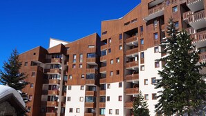 Um edifício de vários andares com varandas, uma pista de esqui ao fundo e neve no chão.