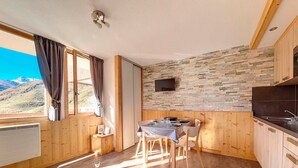 Um quarto aconchegante com uma mesa de jantar posta para dois, uma TV fixada em uma parede de pedra, e vista para montanhas pela janela.