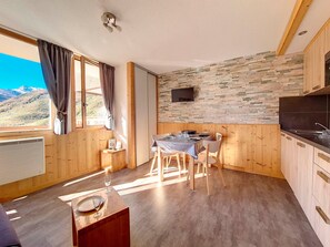 A cozy room with a dining table set for two, a TV mounted on a stone wall, and a view of mountains through a window.