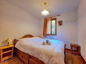 Ein Schlafzimmer mit einem hölzernen Bett, einem Nachttisch mit Lampe, einem Fenster mit Vorhängen und einem kleinen Bild an der Wand.
