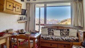 Une pièce chaleureuse avec un bureau en bois, un lit avec une literie à motifs, une fenêtre donnant sur les montagnes et un ventilateur de plafond.
