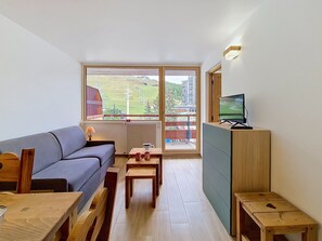 A compact living space with a sofa, a wooden table, a television on a cabinet, and a view of a balcony with a rural landscape.