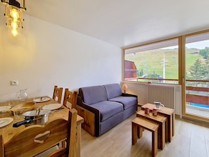 A room with a dining table, wooden chairs, a sofa, and a view of a green landscape through a large window.