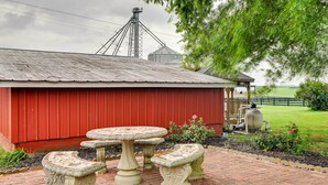 Ein rotes Backstein-Stallgebäude mit einer Metallbedachung, umgeben von einer Ziegelterrasse mit Steintischen und -b änken.