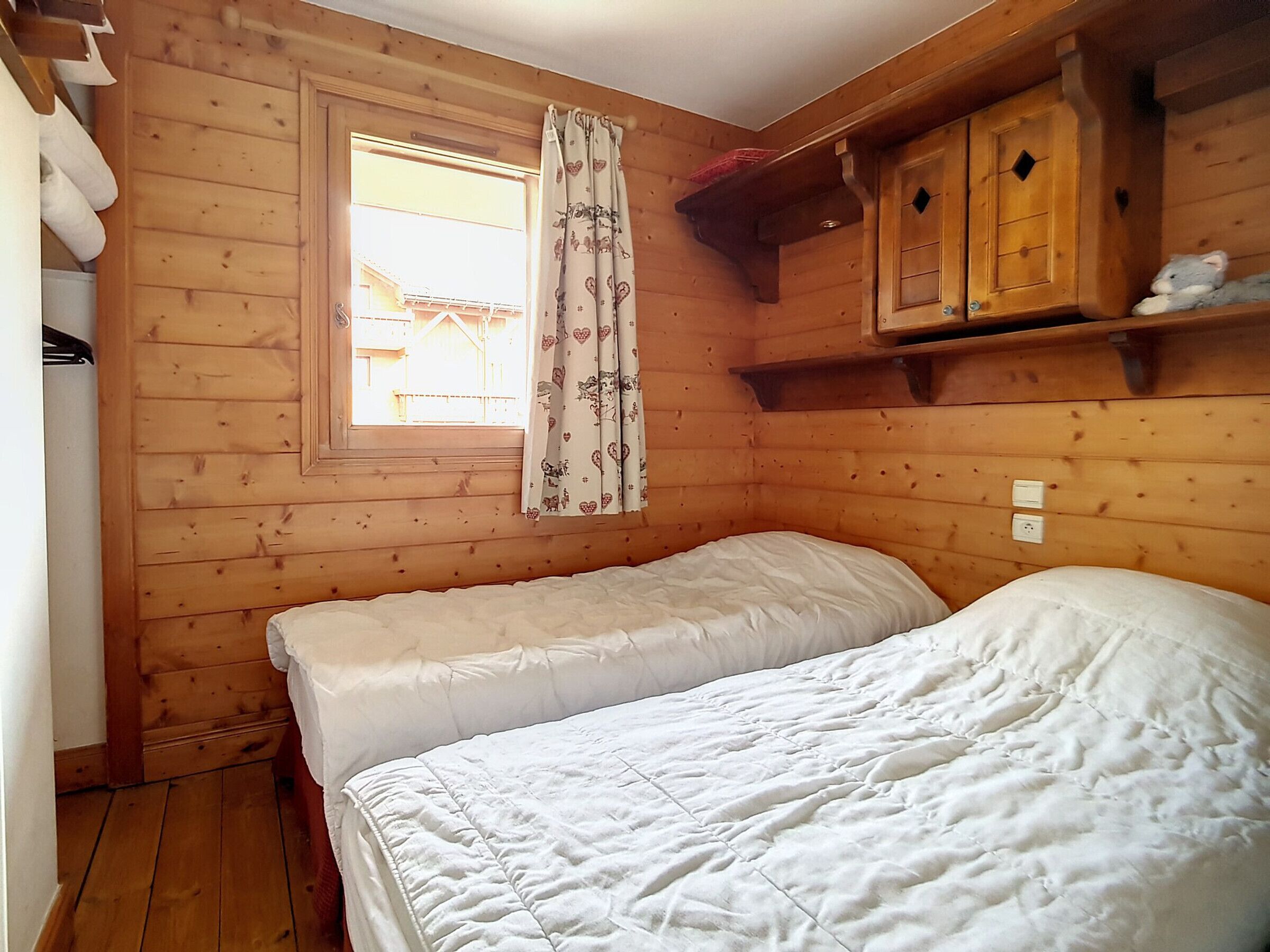 Une chambre dans un chalet en bois, avec deux lits, une fenêtre avec des rideaux et une armoire en bois au-dessus des lits.