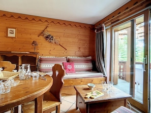 Um interior em madeira com uma mesa de jantar, cadeiras, um banco com almofadas estampadas e vista através de uma porta de vidro.