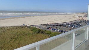 Une vue sur la plage depuis un balcon, avec un parking en contrebas et un ciel dégagé.