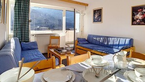 A cozy room with a blue sofa, a wooden table set for dining, and a view of a mountainous landscape through a window. - Ski-in apartment, 2 rooms, 6 ppl, south/west balcony (Les Menuires)
