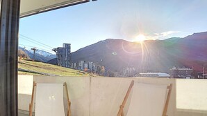 A balcony with two white chairs, a cityscape with mountains in the background, and a bright sun setting or rising. - Ski-in apartment, 2 rooms, 6 ppl, south/west balcony (Les Menuires)