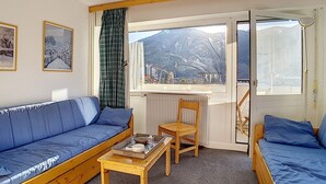 A cozy room with a blue sofa, a wooden table with a tray, a chair, and a view of mountains through a window. - Ski-in apartment, 2 rooms, 6 ppl, south/west balcony (Les Menuires)