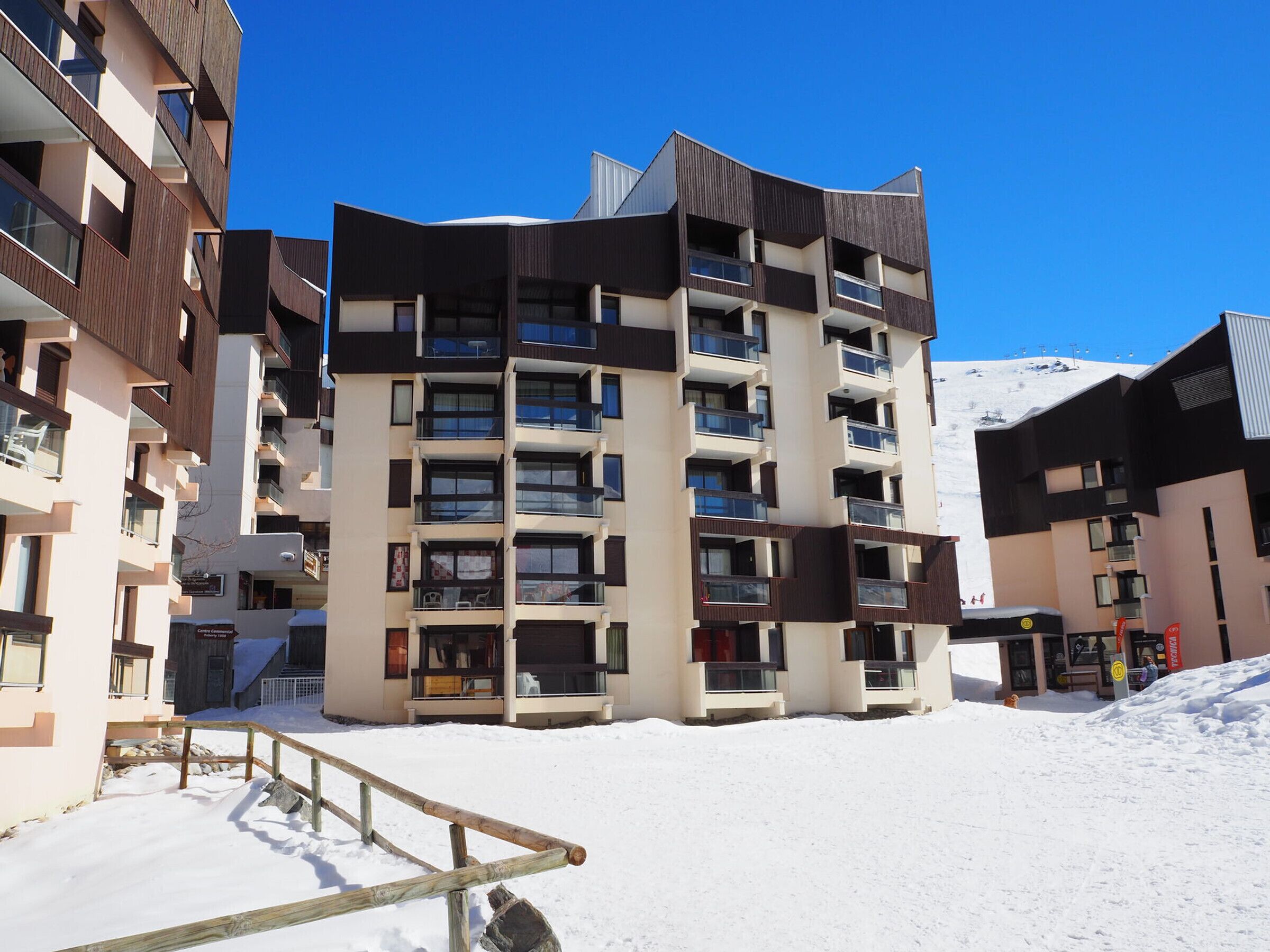 A multi-story hotel with balconies, a ski slope in the background, and a wooden staircase leading to the entrance.