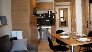 Uma cozinha compacta com eletrodomésticos modernos, um espaço de jantar com mesa de madeira posta para dois e uma área de estar acolhedora com sofá e mesa de centro.
