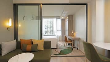 A modern hotel room with a green sofa, a white round table, a desk, and a view of a cityscape through a large window.