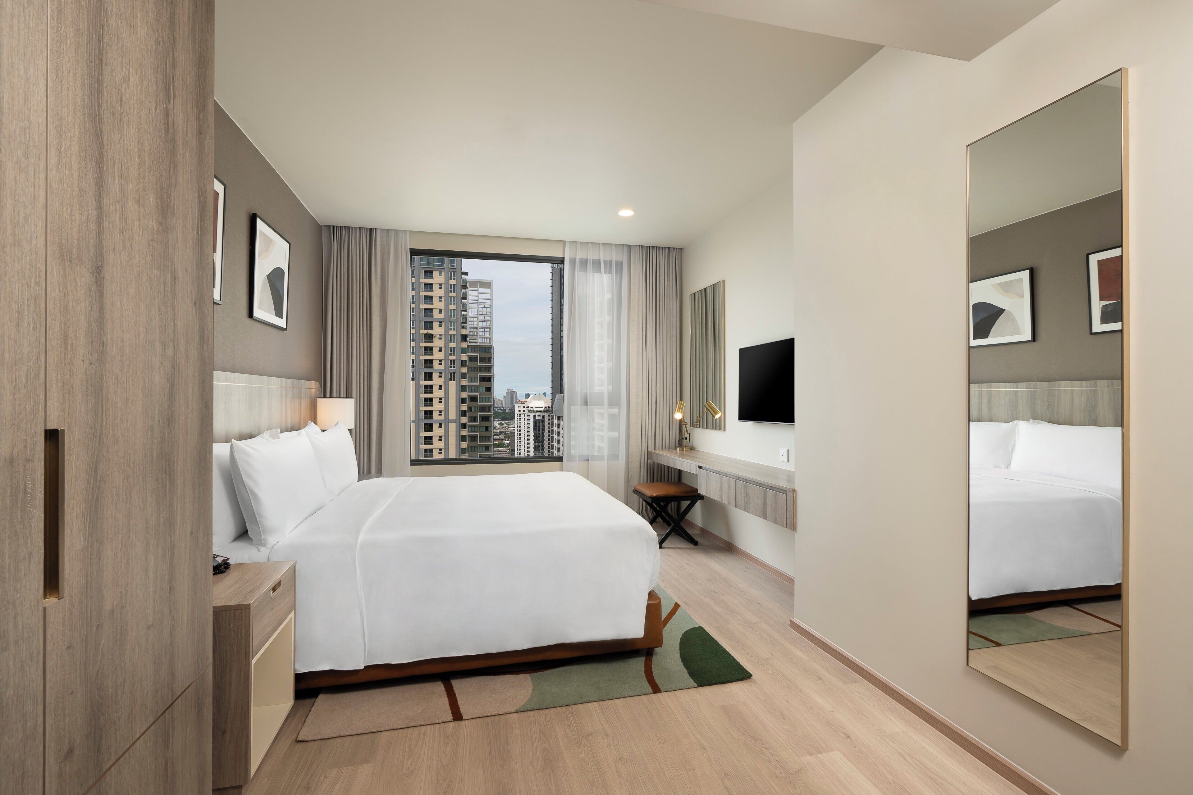 A modern hotel room with a large bed, a desk, a chair, a TV, and a view of the city through large windows.