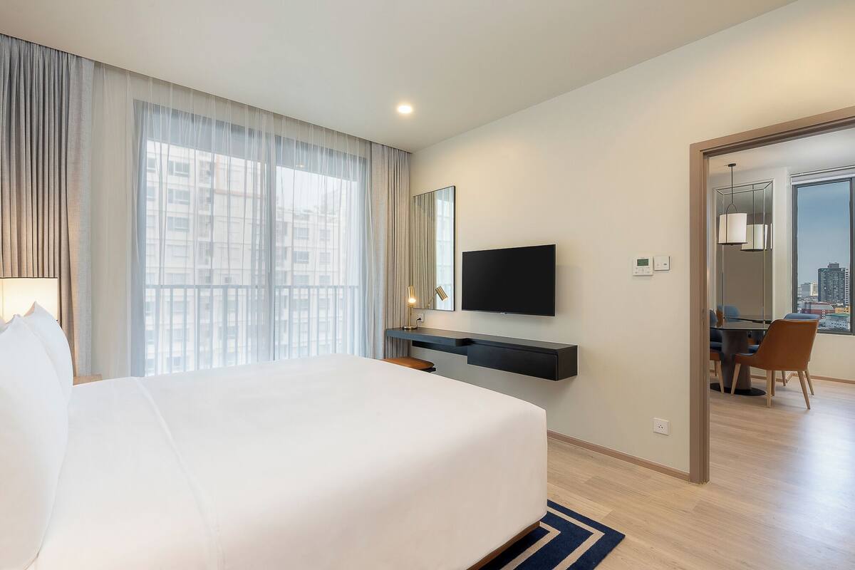 Une chambre d’hôtel avec un grand lit, un téléviseur fixé au mur, un bureau avec une chaise et une vue sur la ville par la fenêtre.