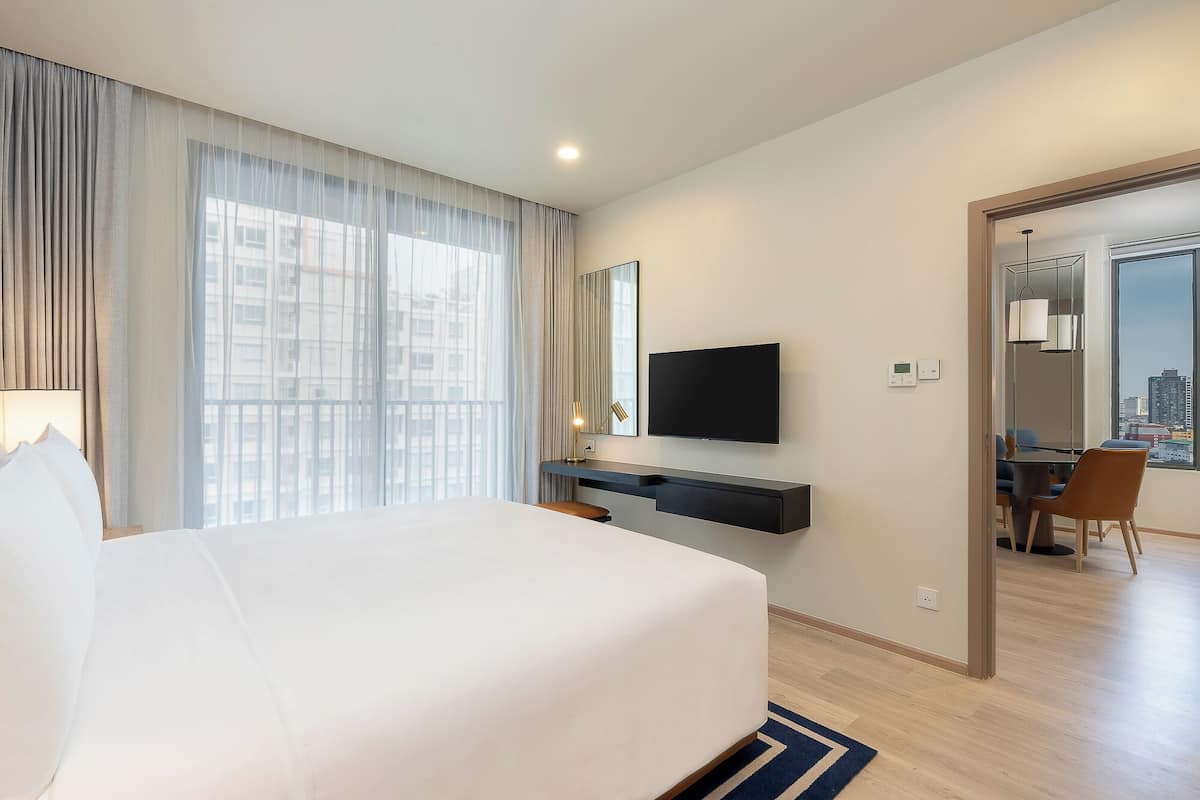 Habitación de hotel con una cama grande, televisor montado en la pared, un escritorio con silla y vista a la ciudad por la ventana.