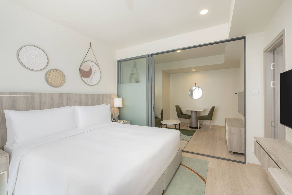 Une chambre d’hôtel dotée d’un grand lit, d’un bureau, d’une chaise, d’une télévision et d’un coin repas visible à travers une porte vitrée.