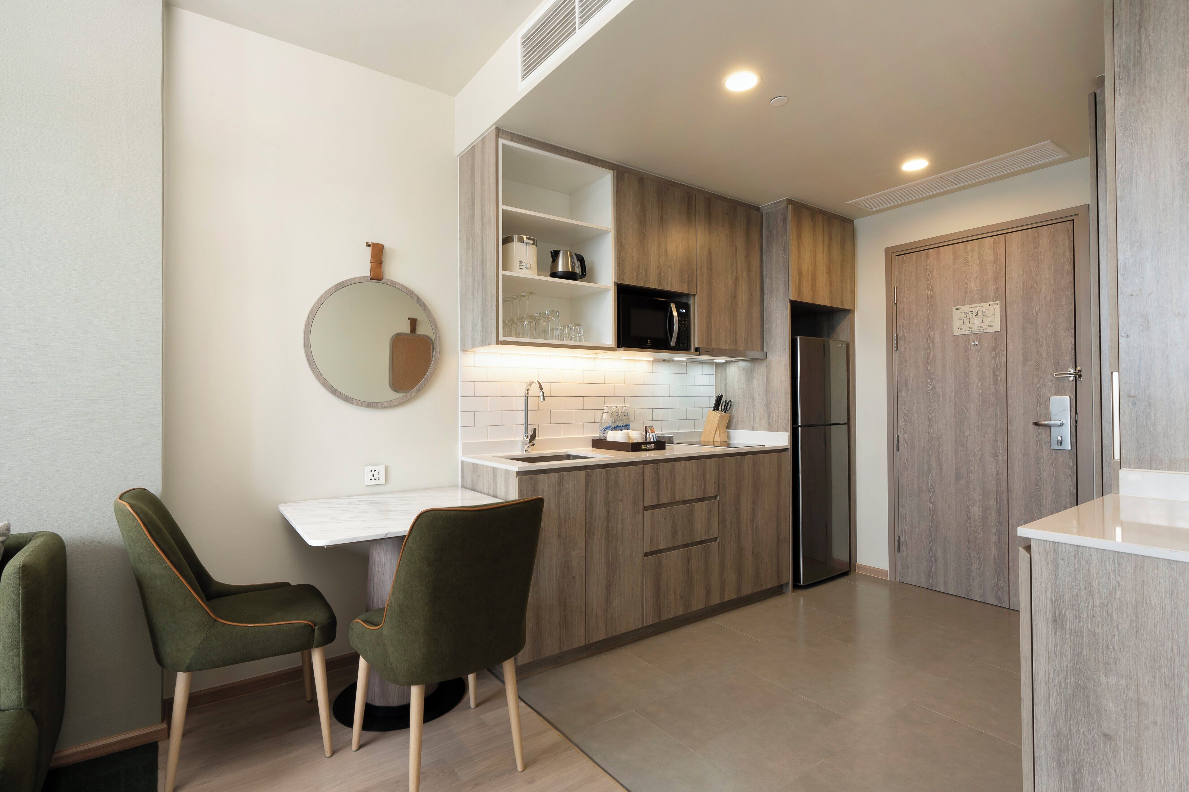 A modern kitchen with wooden cabinets, a built-in microwave, and a refrigerator. There are two green chairs and a white countertop.